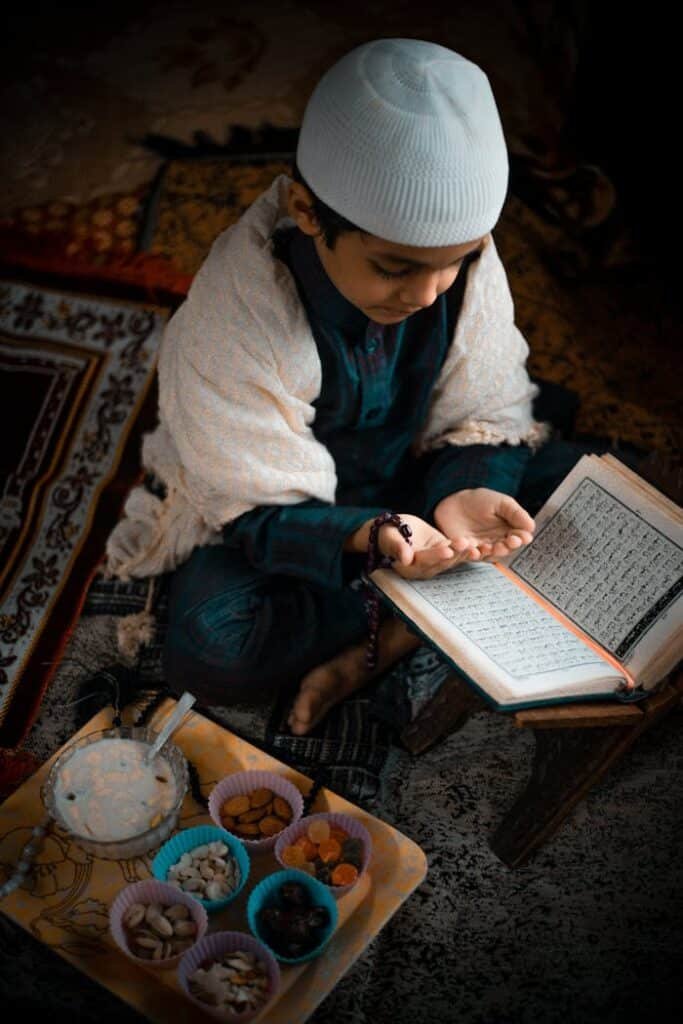 pexels photo 4521955 A young boy in traditional attire reads and prays with an open Quran beside snacks.