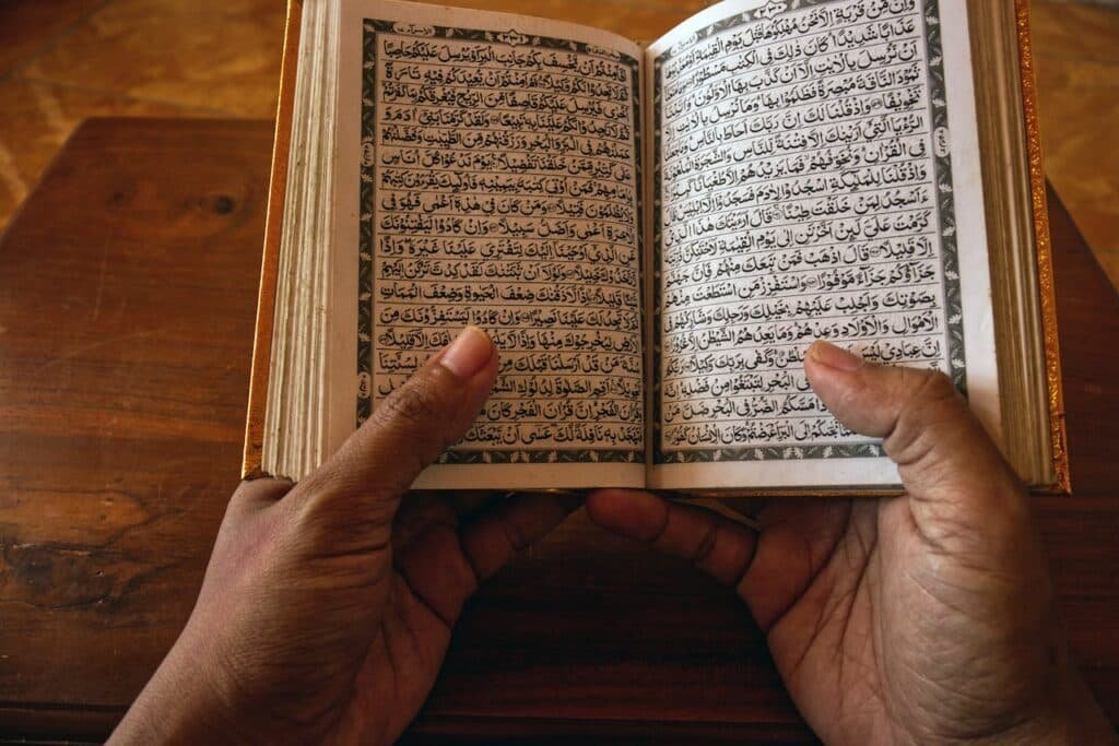 pexels photo 7261978 A close-up of hands holding an open Quran on a wooden table, showcasing Arabic script.